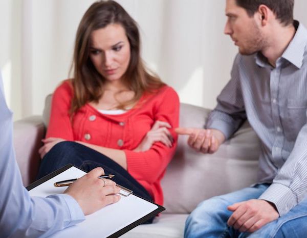 man and woman sitting on couch talking to therapist holding clipboard